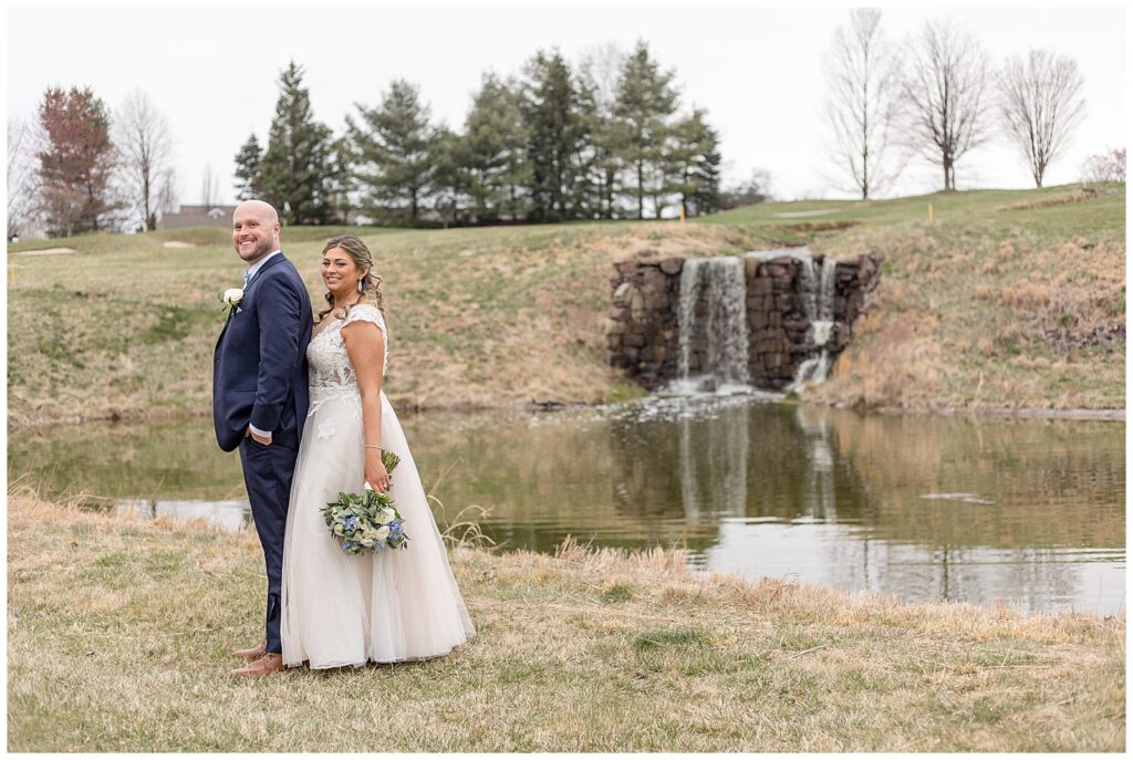 bride standing behind her groom by pond with small waterfall at rivercrest golf club on spring wedding day
