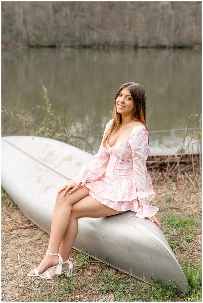 senior girl in pastel pink and white dress sitting on top of silver canoe by speedwell forge lake in lititz pennsylvania