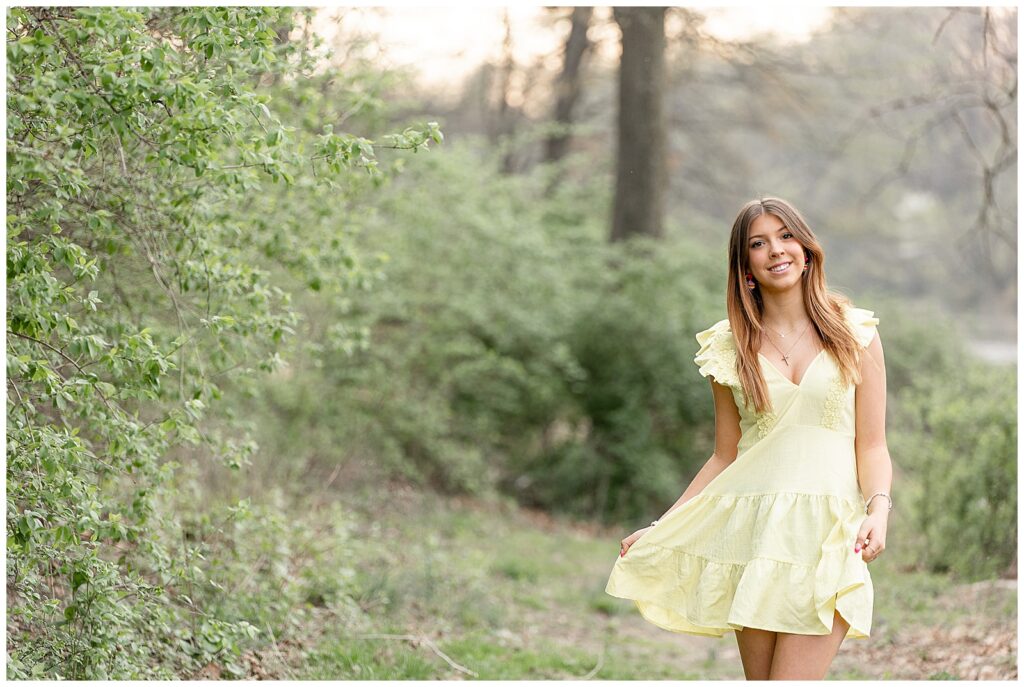 senior girl flaring our edge of her yellow dress with bright smile  by trees at speedwell forge lake in lancaster pa