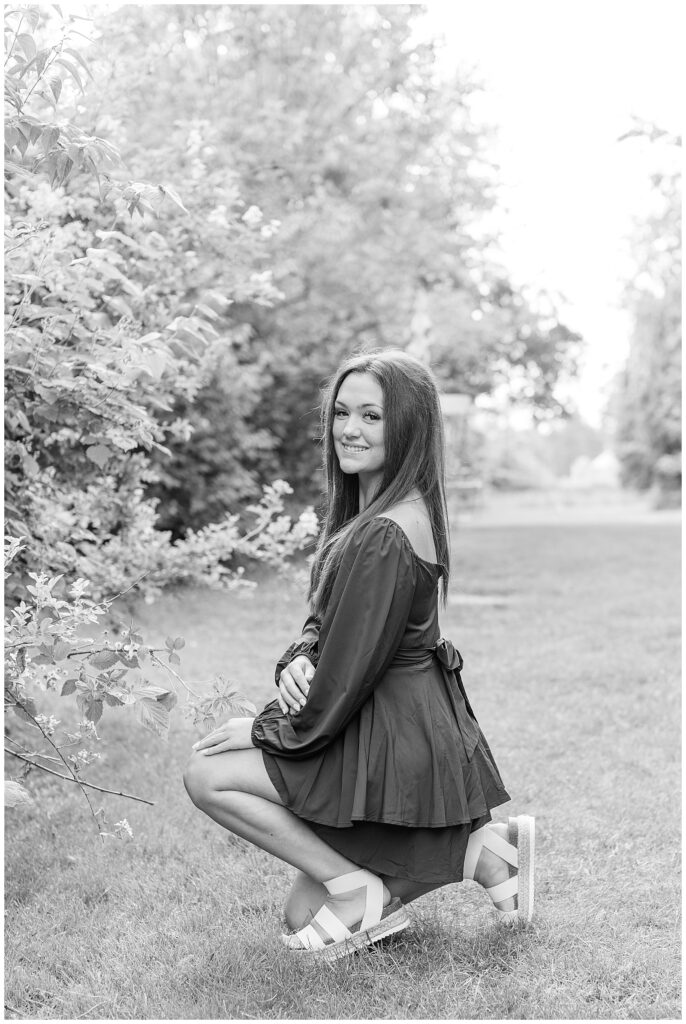 black and white photo of senior girl crouching down in grass by large bush with left shoulder toward camera at overlook park