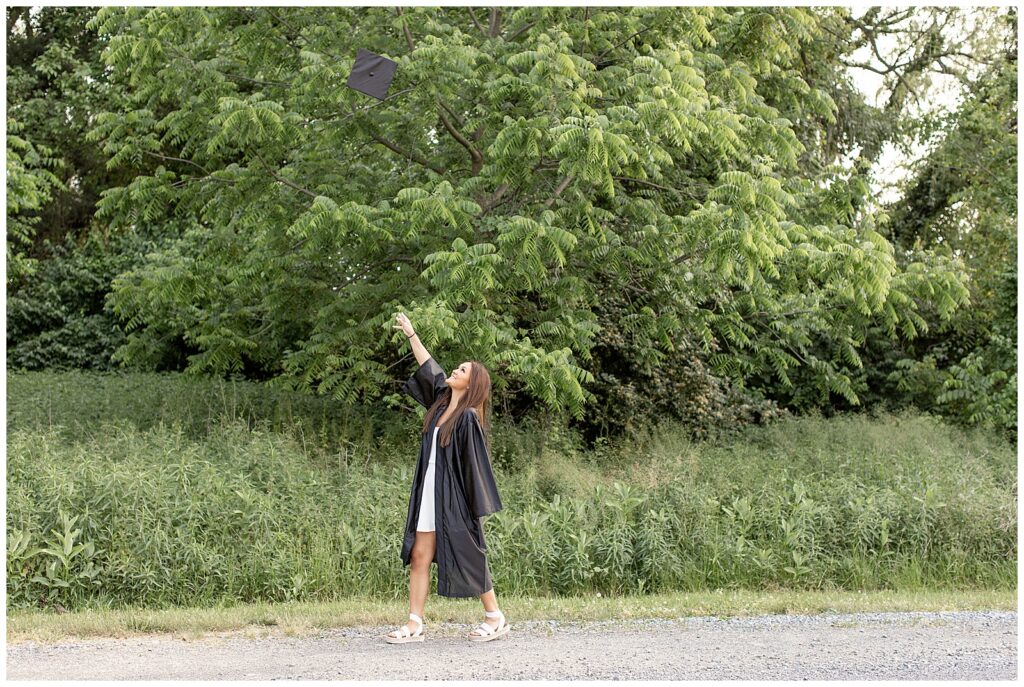 senior girl in unzipped black graduation gown with white dress showing walking along path with right arm in the sky at park in manheim township