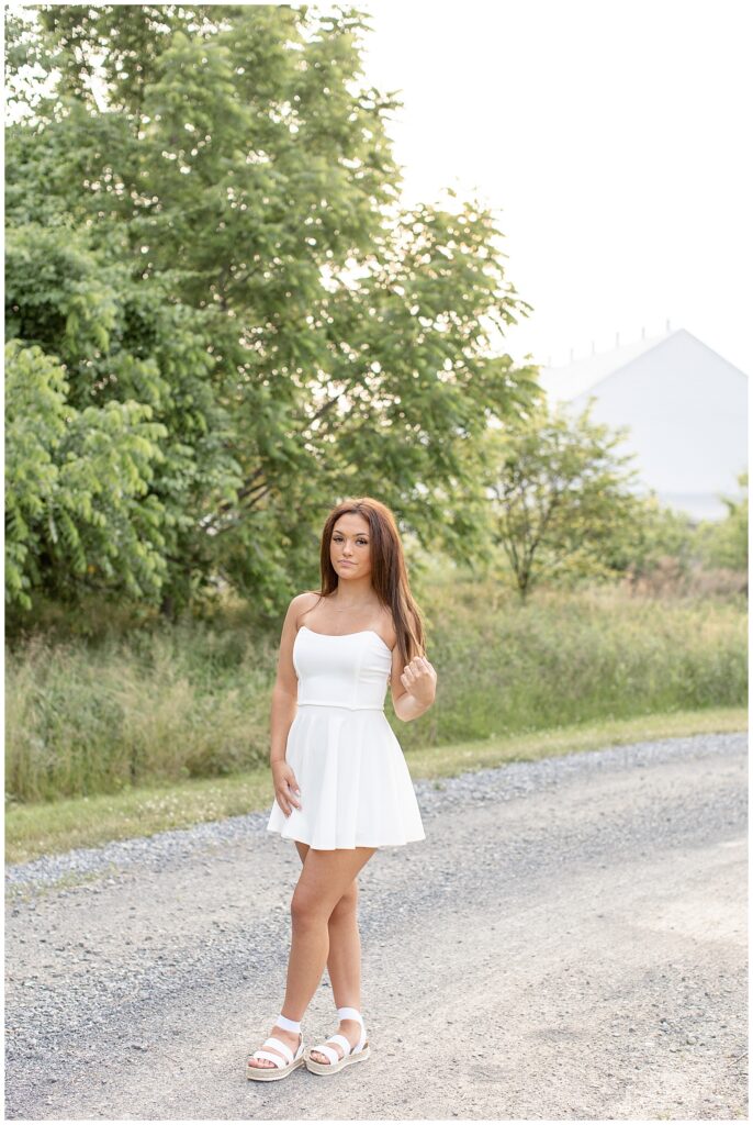 senior girl in white dress walking along path holding onto edge of her long hair with left fingers on sunny evening at park in lancaster county