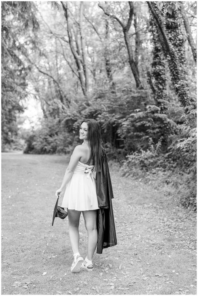 black and white photo of senior girl walking away from camera and looking back over left shoulder with her graduation gown slung over her right shoulder in lancaster pennsylvania