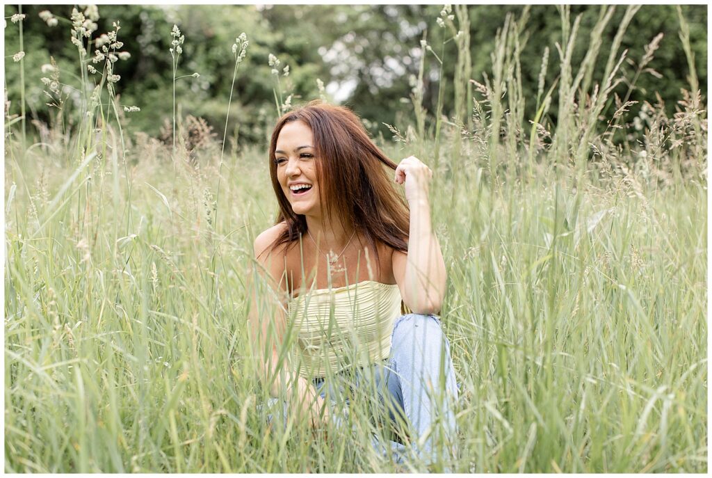 senior girl sitting in tall wild grasses and looking to her right smiling as she holds edge of her hair with left hand at overlook park
