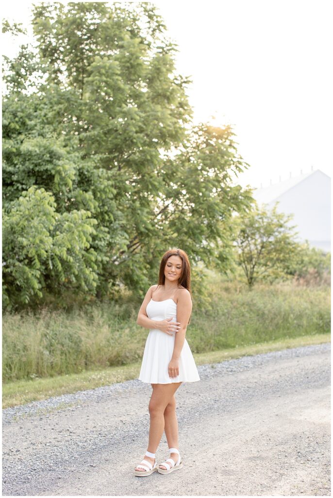 senior girl in white dress standing in pathway with her right hand on her left elbow and looking down on spring evening at overlook park
