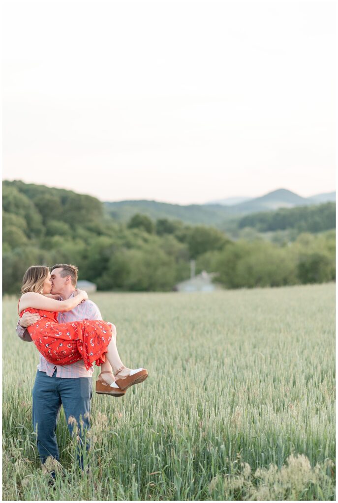 man holding his woman in his arms off the ground as they kiss by wheat field and mountains in pennsylvania