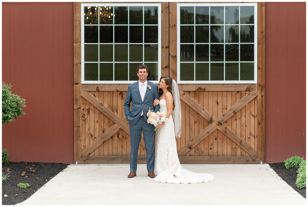 groom smiles at camera as bride holds onto his left arm and looks up at him smiling by barn door at promise farm