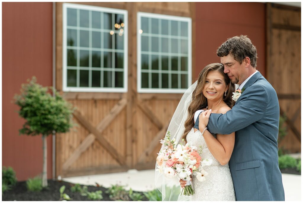 groom hugs his bride from behind as she smiles at camera by barn venue at promise farm