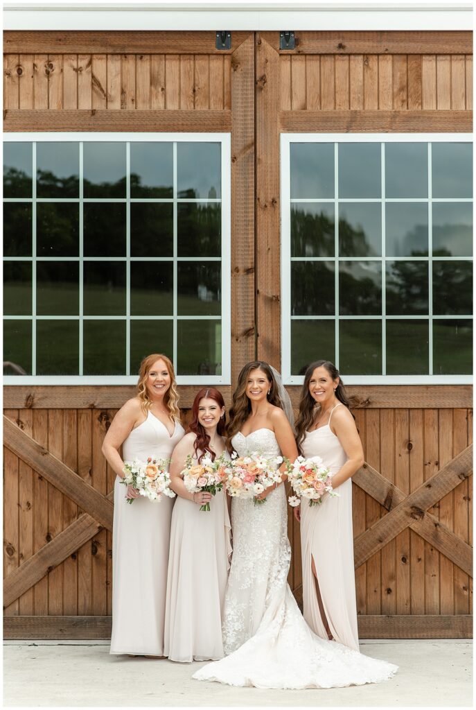 bride with her three bridesmaids all wearing beige gowns holding bouquets by barn doors at promise farm
