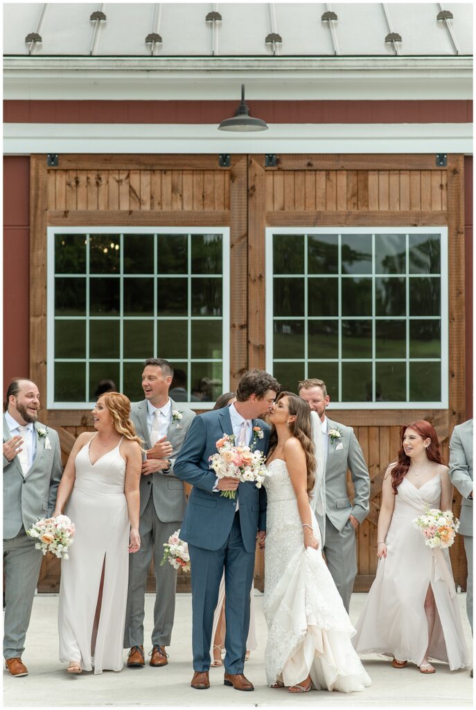 couple kissing as their bridal party is casually walking and talking behind them by wooden doors at promise farm