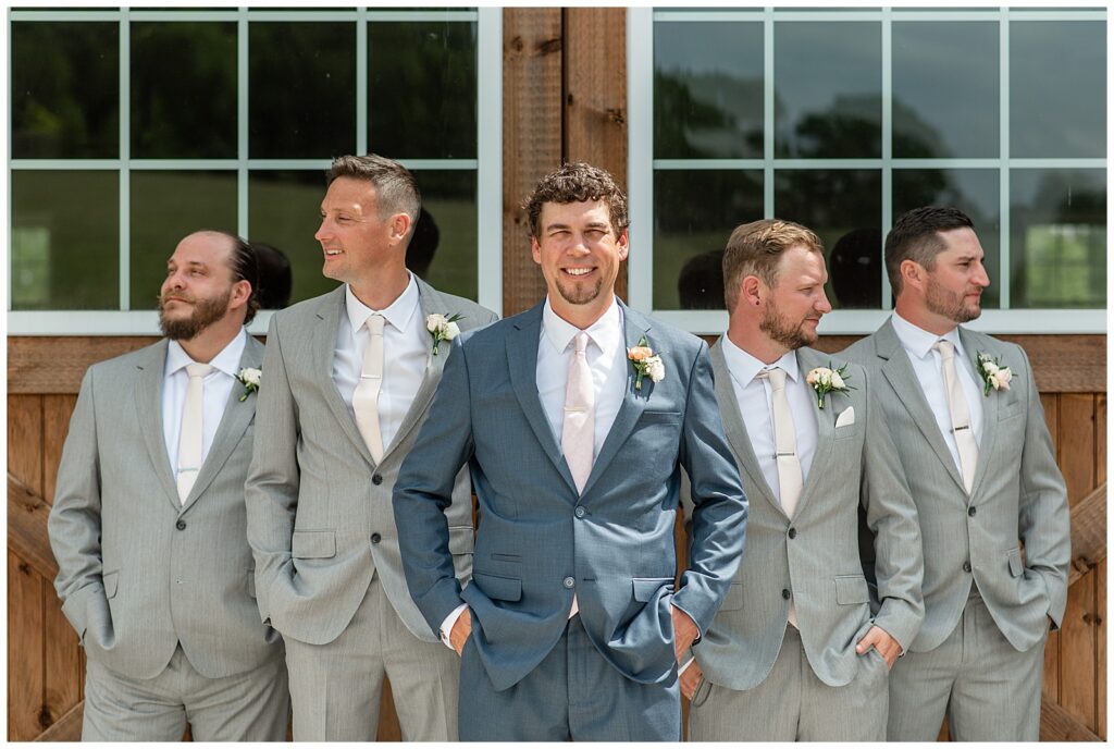 groom wearing navy blue suit standing in front of his four groomsmen wearing gray suits by barn door at promise farm