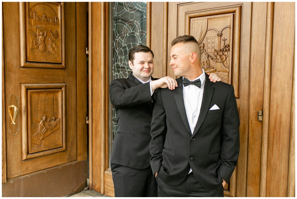 best man resting his hands on groom's shoulders as groom looks back at him and they both wear black tuxes outside Saint Ignatius Catholic Church