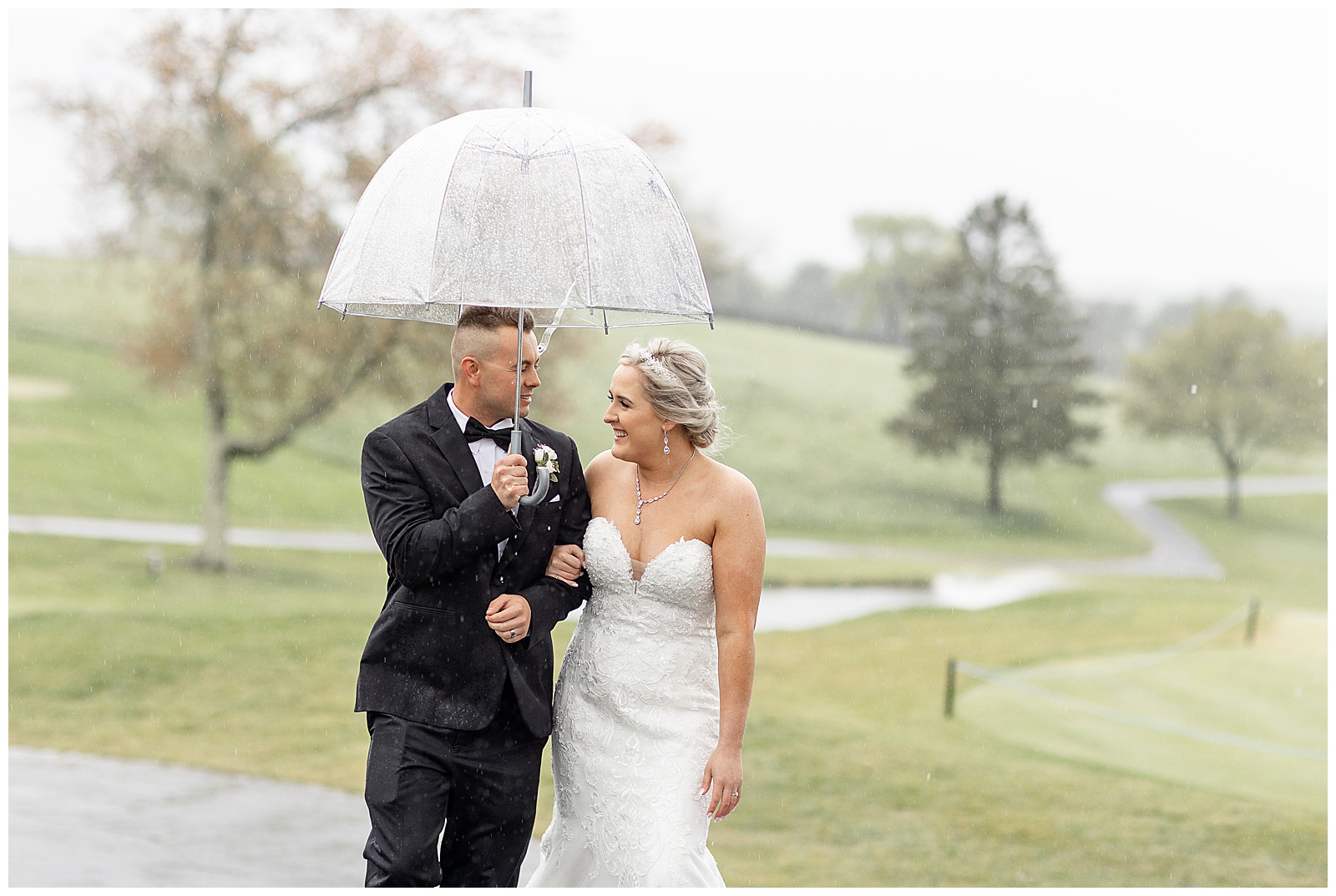 bride and groom huddle under cute clear umbrella on golf cart path on rainy wedding day in maryland