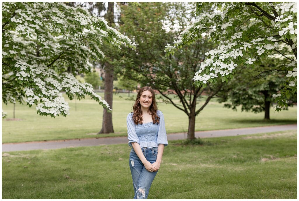 senior girl with left ankle crossed over right one smiling with her hands held in front of her by blooming white trees at the reading public library