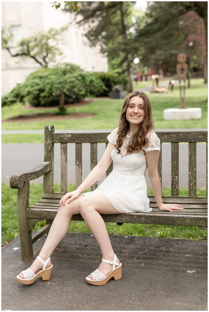 senior girl wearing ivory lace dress sitting on wooden bench and leaning on left hand at outdoors at the reading public library in berks county