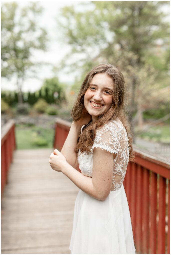 senior girl smiling and holding her right elbow with her left hand by red wooden bridge outdoors in berks county pennsylvania