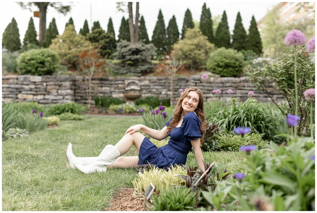 senior girl wearing dark blue dress and white cowboy boots sitting grass with legs extended and looking back over left shoulder at the reading public library