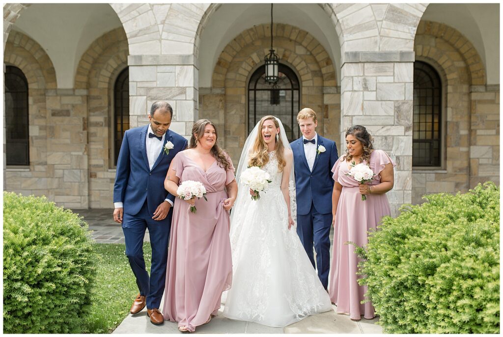 bride with their two bridesmaids in pink and two groomsmaids in blue by stone archway at the shrine of saint anthony in maryland