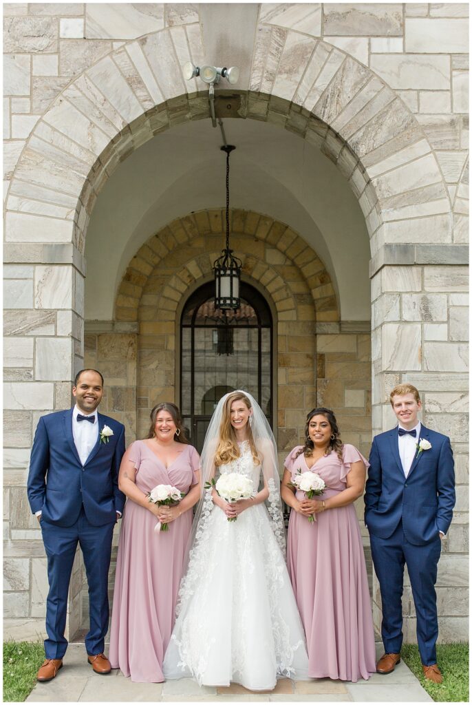bride with her two bridesmaids and her two groomsmaids standing in a row by large archway at the shrine at saint anthony