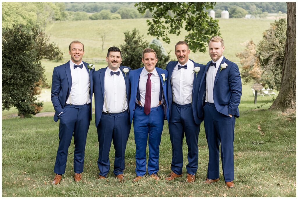 groom with his four groomsmen all in blue suits standing with their arms around one another by field in maryland