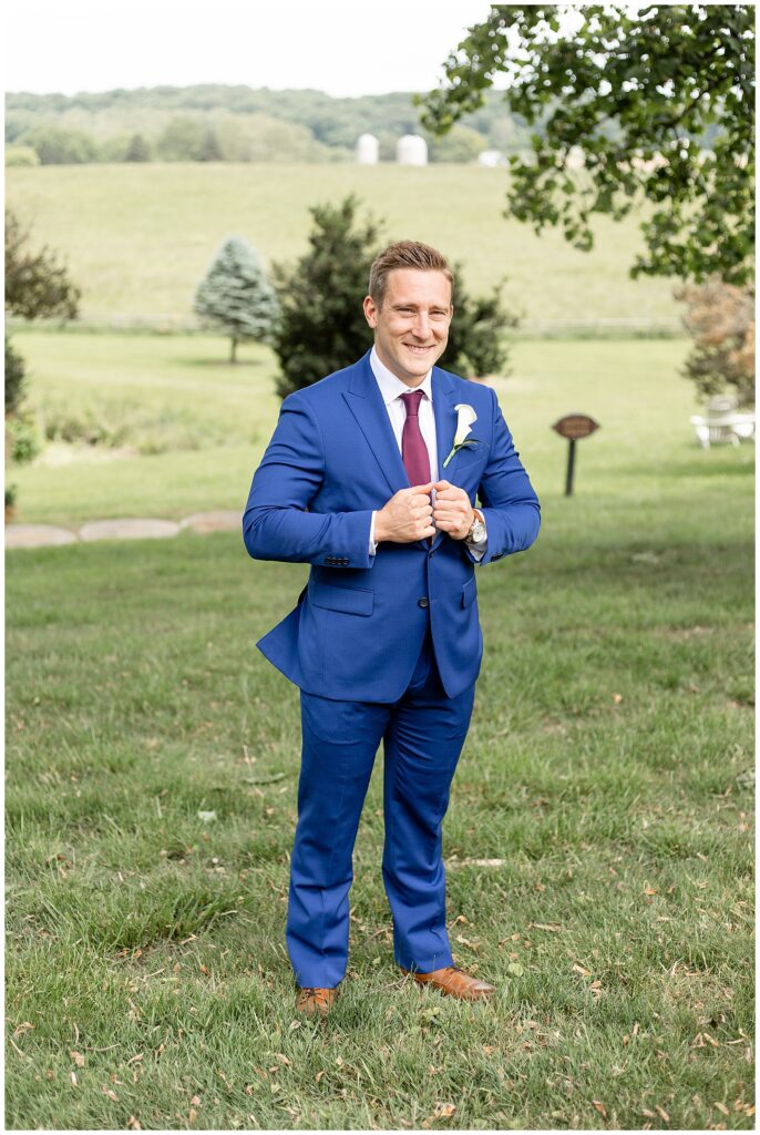 groom holding onto edge of his suit coat and smiling at the camera by field in maryland