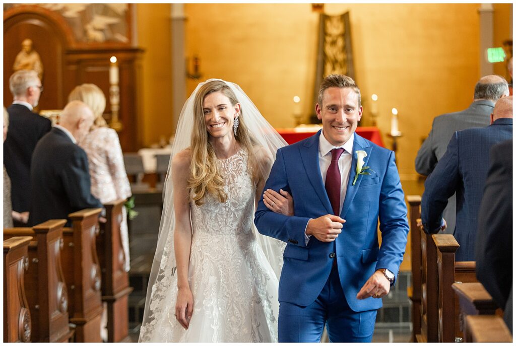 bride and groom with arms linked smiling as they walk down the aisle leaving their wedding ceremony at the shrine of saint anthony