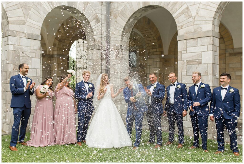 bride and groom surrounded by their bridal party as groom pops confetti outside of the shrine of saint anthony in maryland