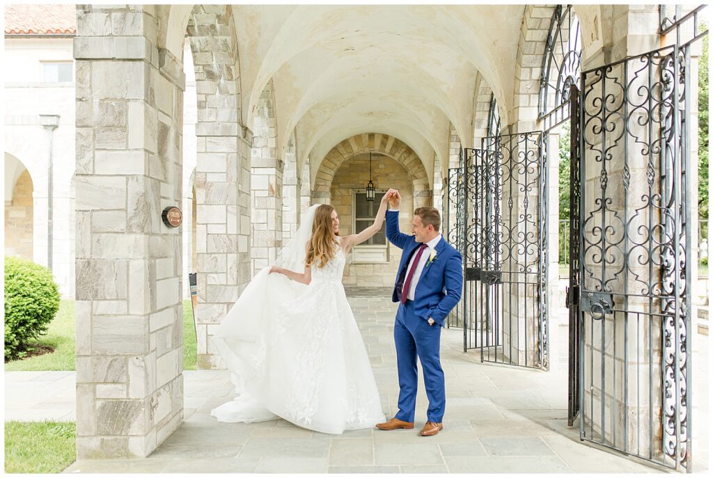 groom twirling his bride under his left arm as she flares out her white dress train by stone archways at catholic church in ellicott city maryland