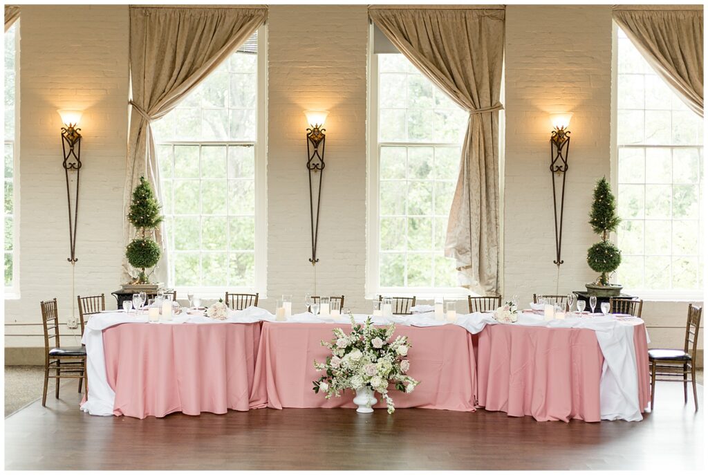 bridal table with light pink linens and large bouquet of white flowers in front of tables at reception at historic savage mill