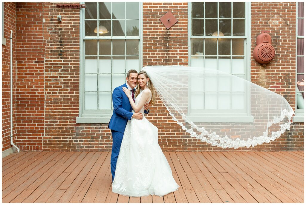 bride and groom standing close with bride's white veil blowing behind them by brick building at historic savage mill