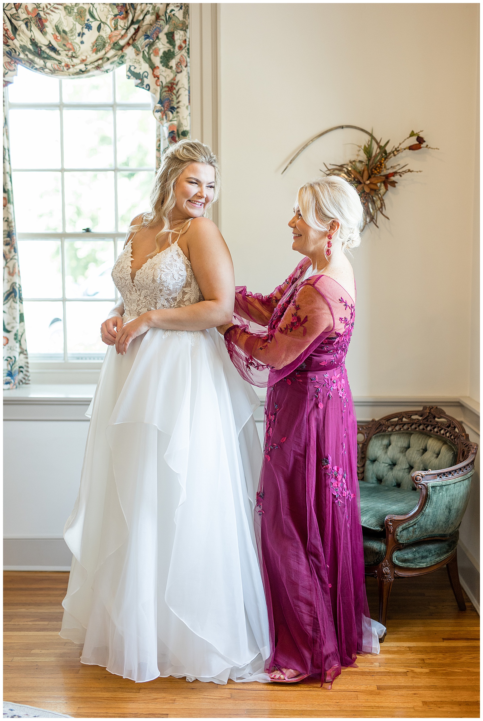 bride looks back over her left shoulder and smiles at her mom helping to zip the back of her dress in bridal suite at the barn at silverstone
