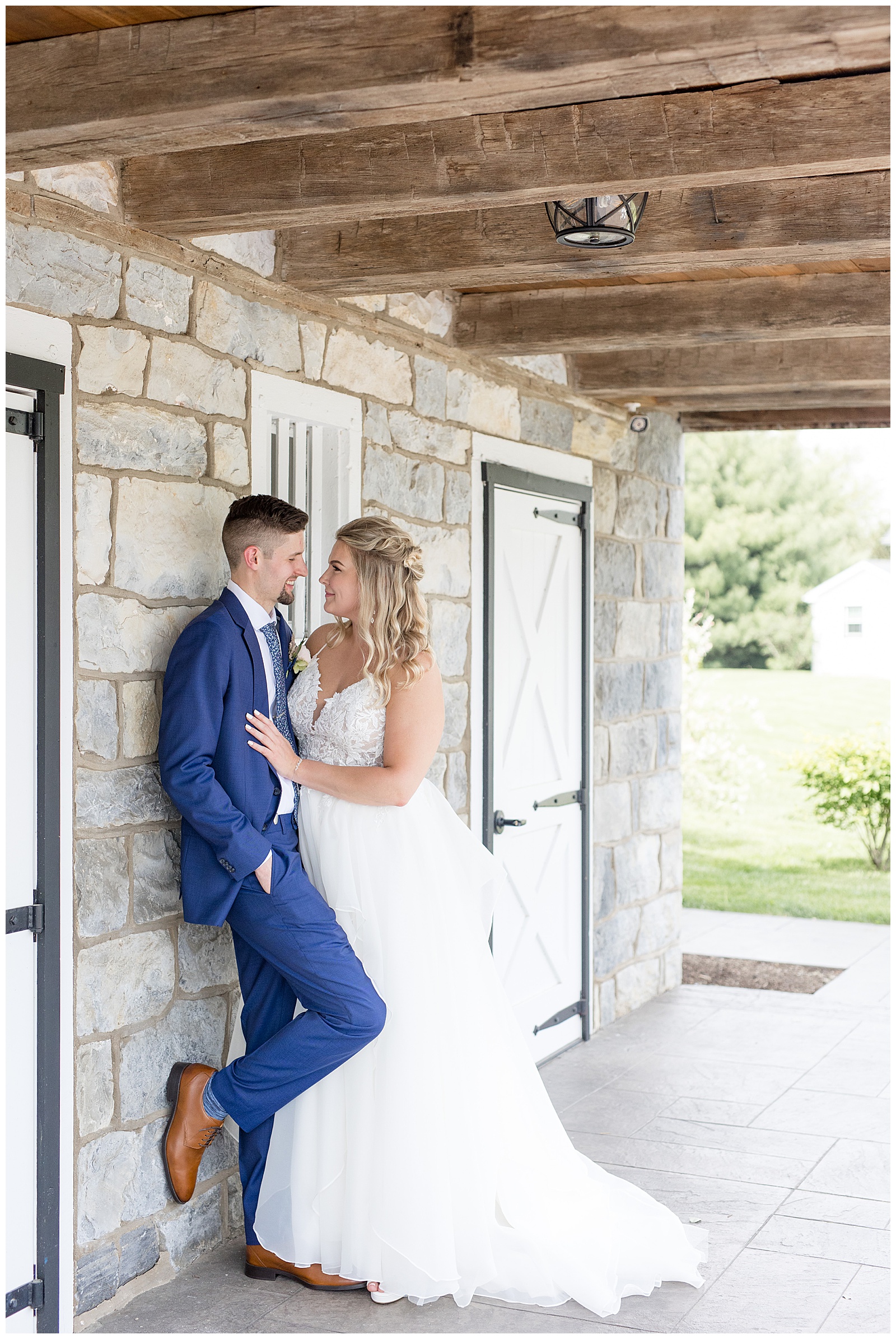 bride leans against her groom as he leans against stone wall of barn at wedding venue in lancaster county