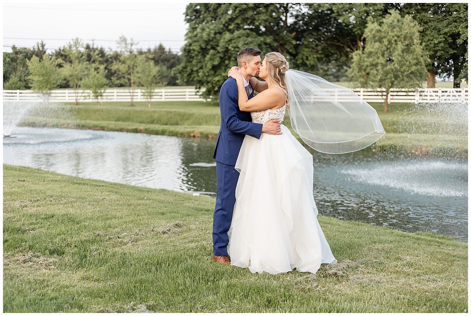 bride and groom kiss by pond as bride's veil blows in the breeze at the barn at silverstone