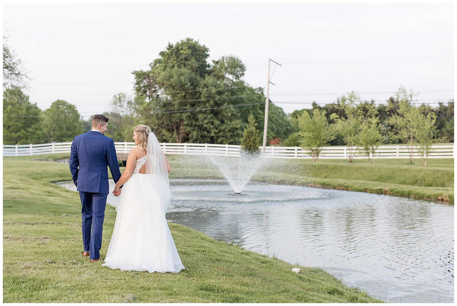 bride and groom hold hands by pond with fountain with their backs toward camera in lancaster pennsylvania