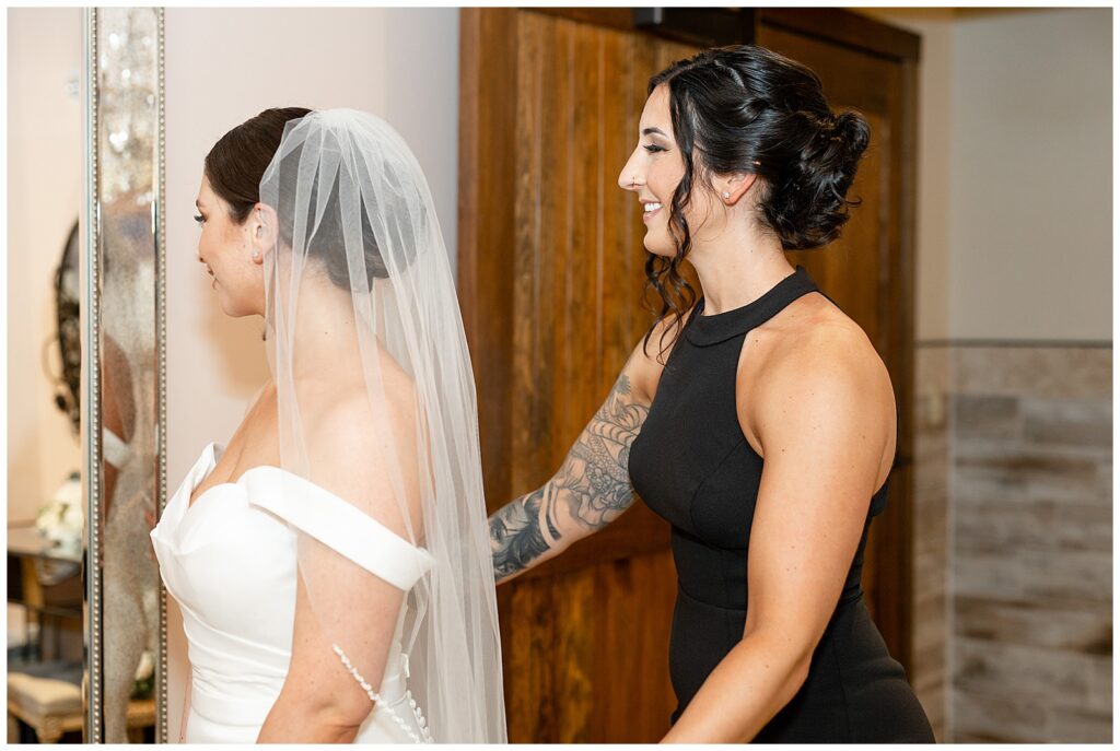 bride in white gown with her matron on honor in black dress smiling as they look in mirror out of view at folino estate