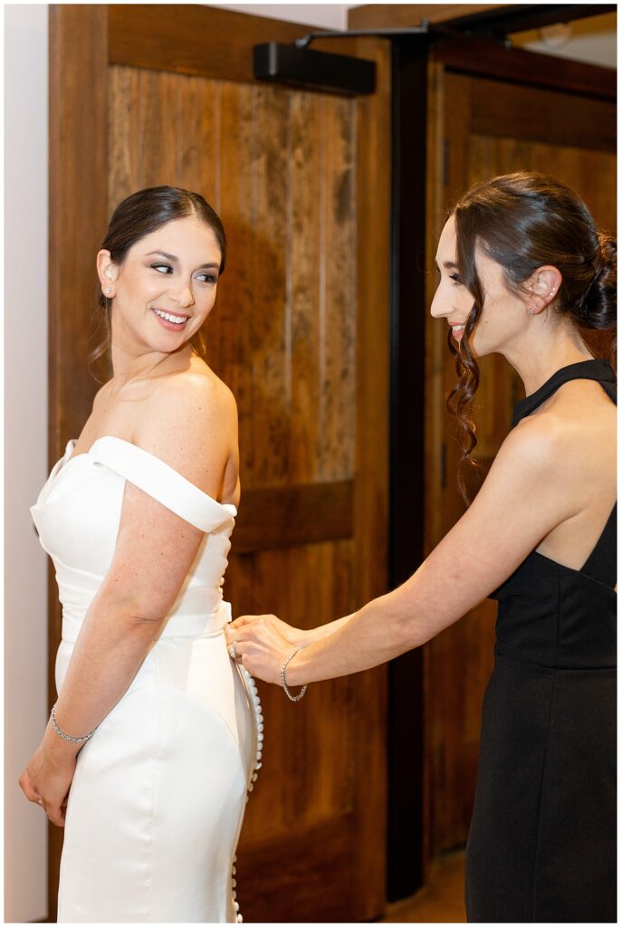 bride looking back over her left shoulder and smiling at her matron of honor buttoning bride's wedding gown in berks county
