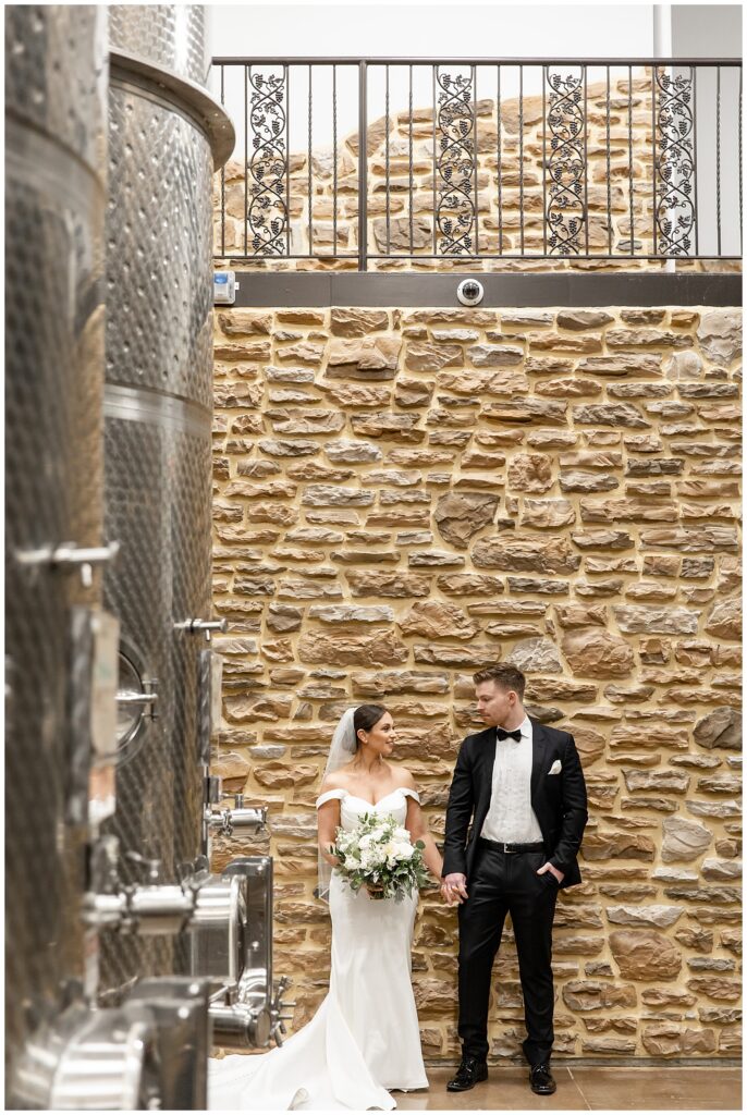 bride and groom hold hands and smile at each other while standing against indoor stone wall at folino estate