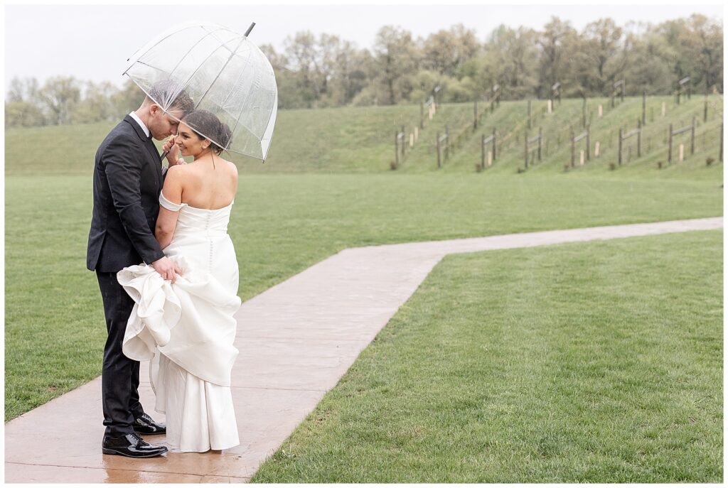 bride and groom nestled under clear umbrella on paved pathway as groom holds bride's dress train at folino estate