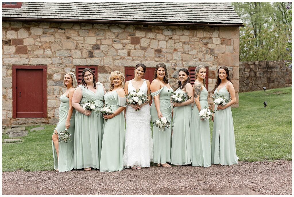 bride in white gown surrounding by her bridesmaids all wearing mint green floor-length gowns by stone building at elizabeth furnace