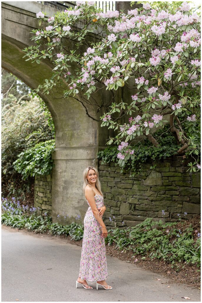 senior girl with right shoulder towards camera standing on paved pathway by large concrete archway at longwood gardens in pennsylvania