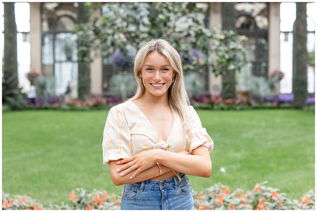 senior girl in beige crop top and blue jeans standing with her arms crossed by her waist and smiling inside arboretum at longwood gardens