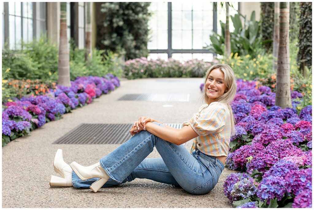 senior girl sitting on floor of arboretum with right leg extended and left knee bent by colorful flowers at longwood gardens