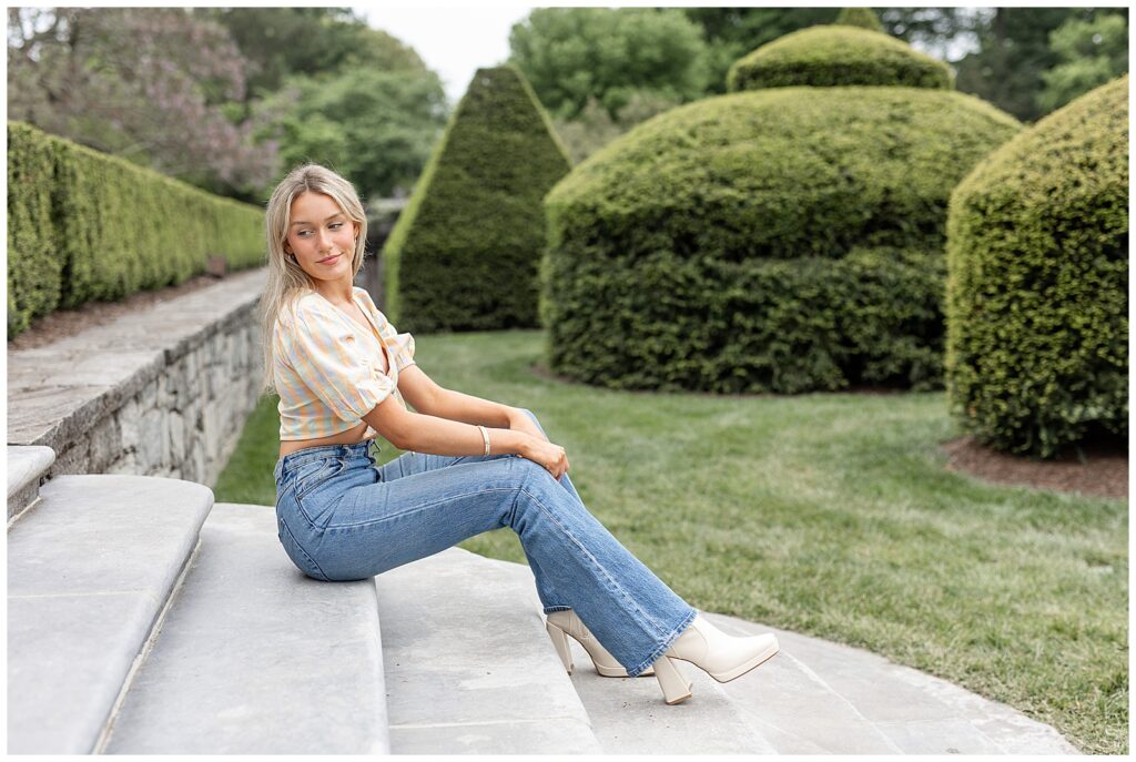 senior girl sitting on concrete steps and looking back over right shoulder with larger bushes in background at longwood gardens