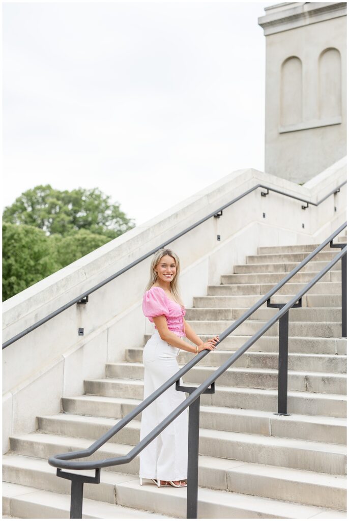 senior girl in pink top and white pants walking up concrete steps holding handrail and looking back at camera at longwood gardens