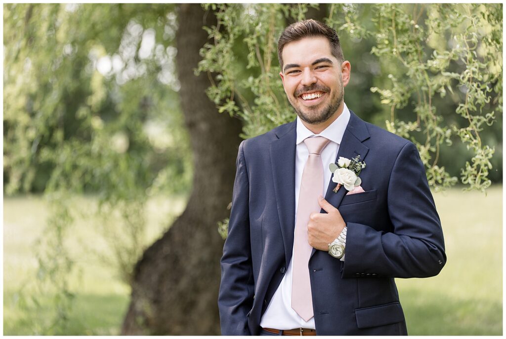 groom smiling as he holds edge of navy suit coat with his left hand by willow tree at the barn at silverstone