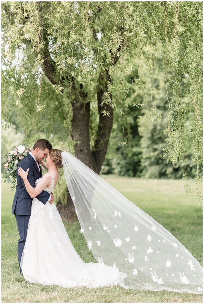 bride and groom touching foreheads together with bride's veil extended behind her by willow tree in lancaster county pennsylvania
