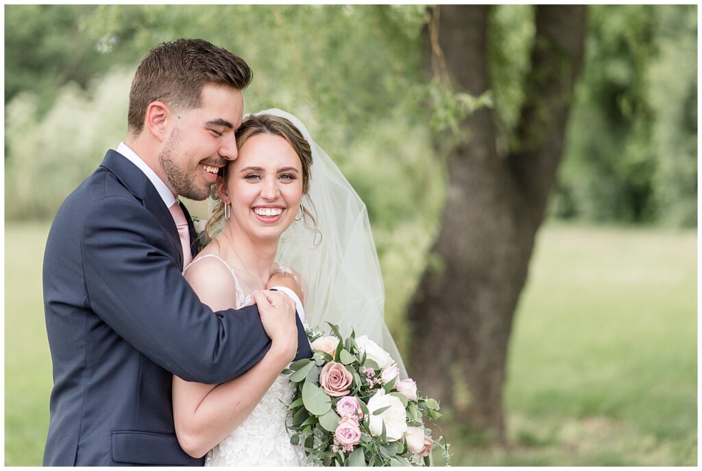 groom hugging his bride from behind as they both smile with willow tree behind them at the barn at silverstone