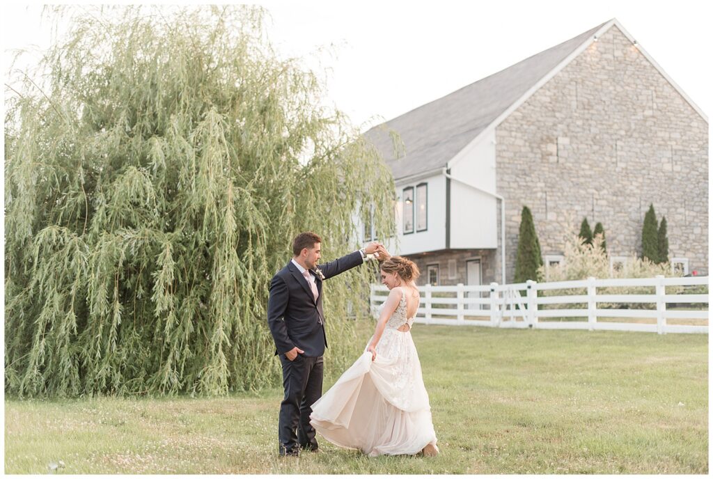 groom twirling his bride under his left hand as she flares out her gown with barn in background at the barn at silverstone