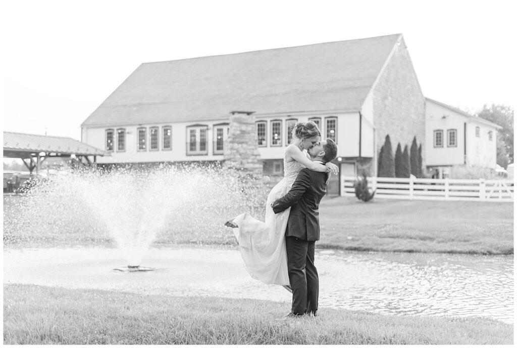 black and white photo of groom lifting his bride off the ground as they kiss by pond at the barn at silverstone