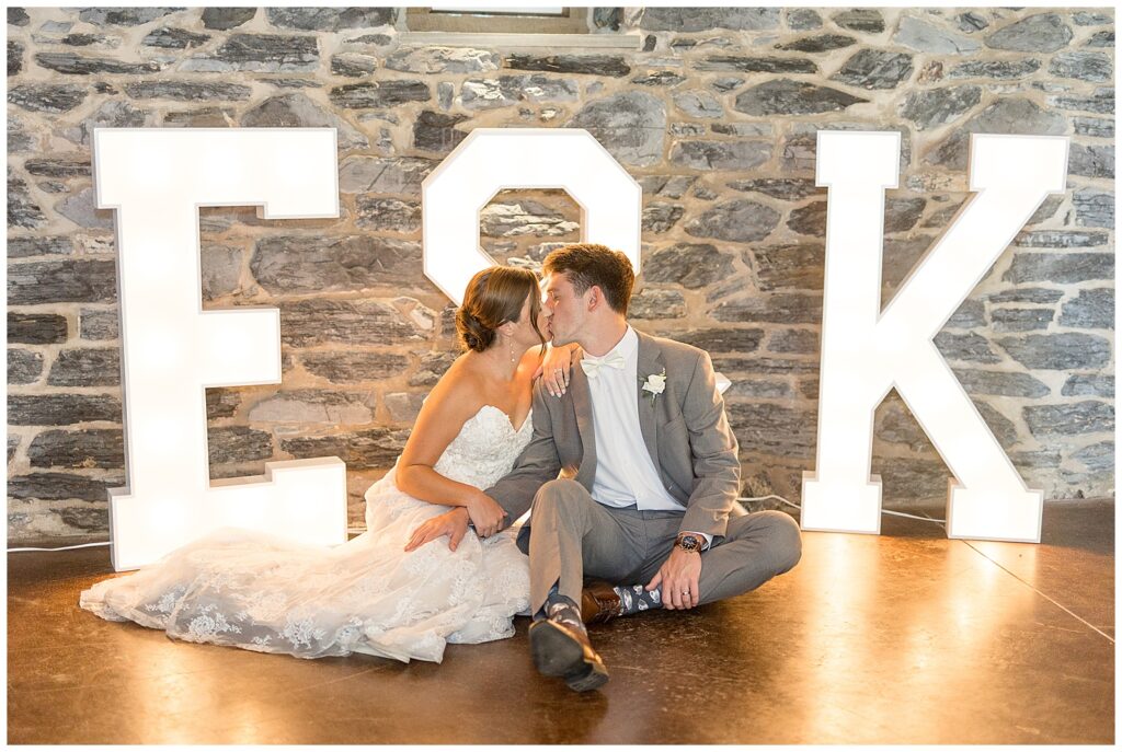 couple sitting on the barn floor kissing with large illuminated E & K behind by stone wall at historic ashland